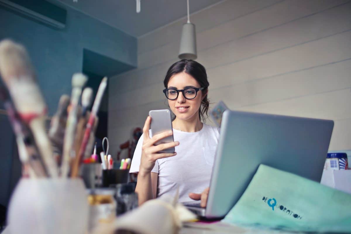 A lady wearing glasses looking at her phone with a laptop open in front of her