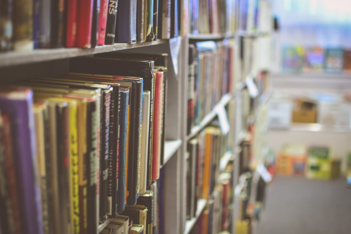 In focus are a range of different books on a shelf, out of focus you can make out the rest of the shelving with books