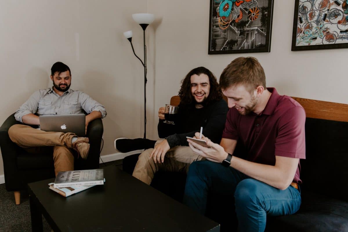 Three men sat on a sofa, one is making notes on a pad and paper, one is using his laptop the other is drinking coffee