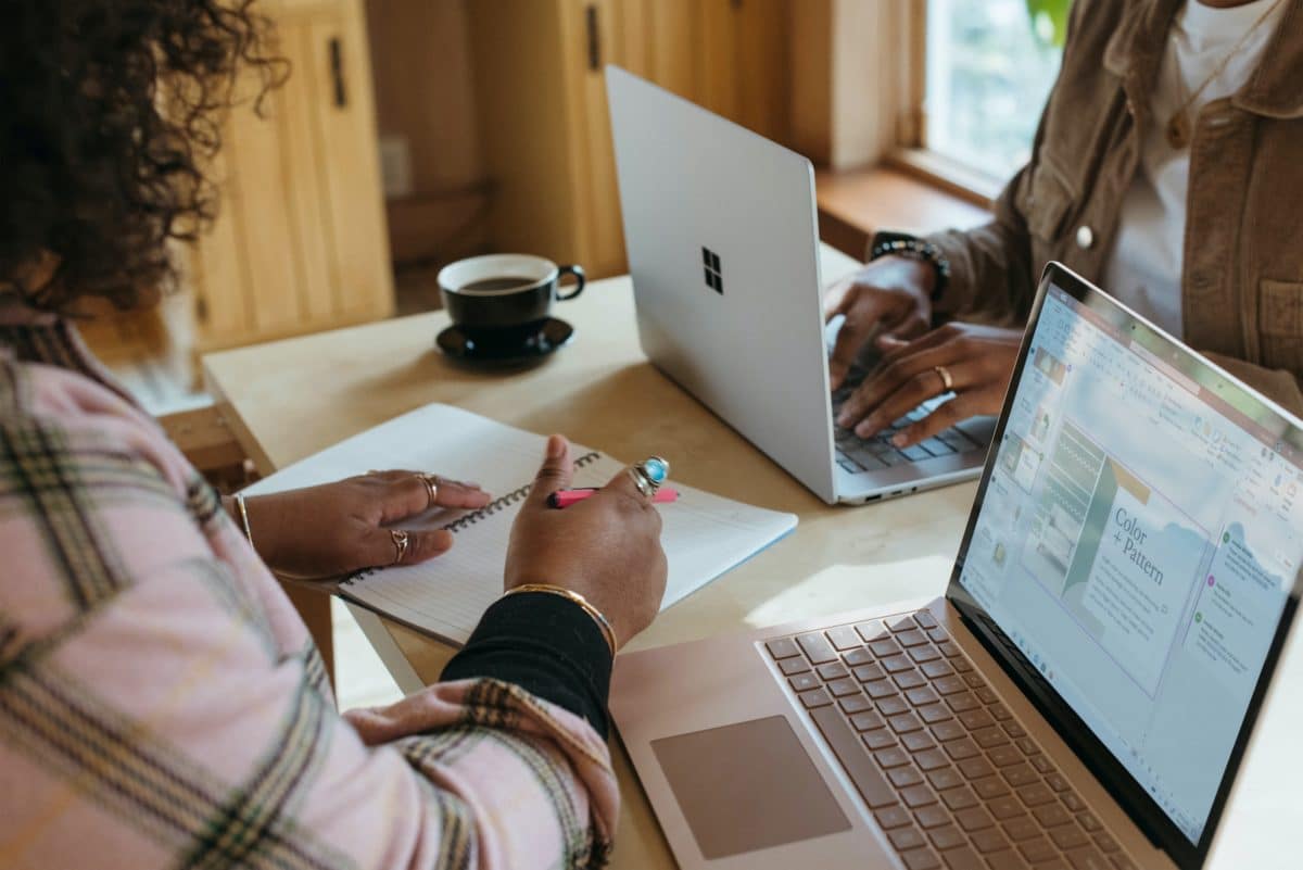 Two people at a desk, one using a laptop the other writing on paper